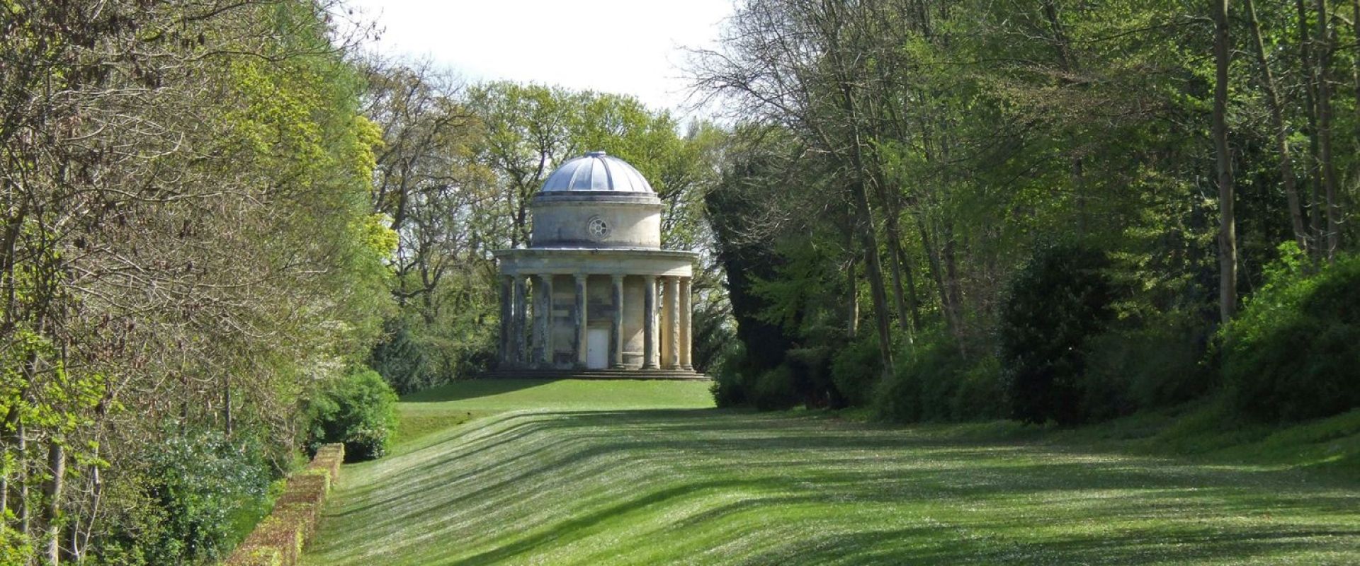 A classical-style rotunda stands at the end of a banked grass promenade lined by trees.