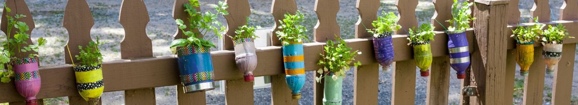 Decorated plastic bottles containing plants hang on a wooden fence.
