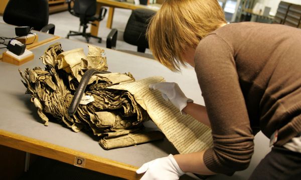 A female archivist wears protective gloves to examine old manuscript records.