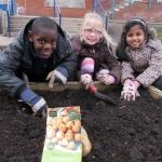 School children pose with a pack of seeds next to an empty raised bed.